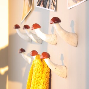 May include: White ceramic mushroom-shaped hooks with red tops mounted on a wall. A yellow knitted scarf is hanging on one of the hooks.