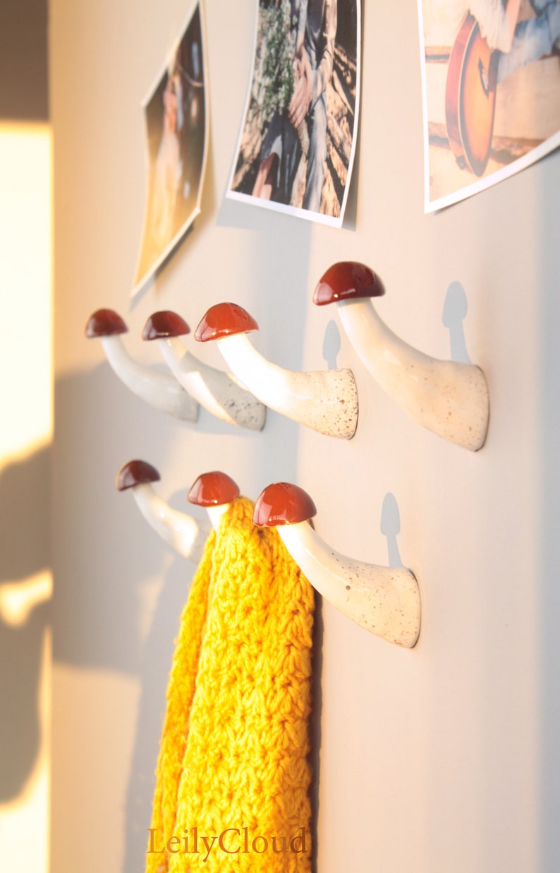 May include: White ceramic mushroom-shaped hooks with red tops mounted on a wall. A yellow knitted scarf is hanging on one of the hooks.