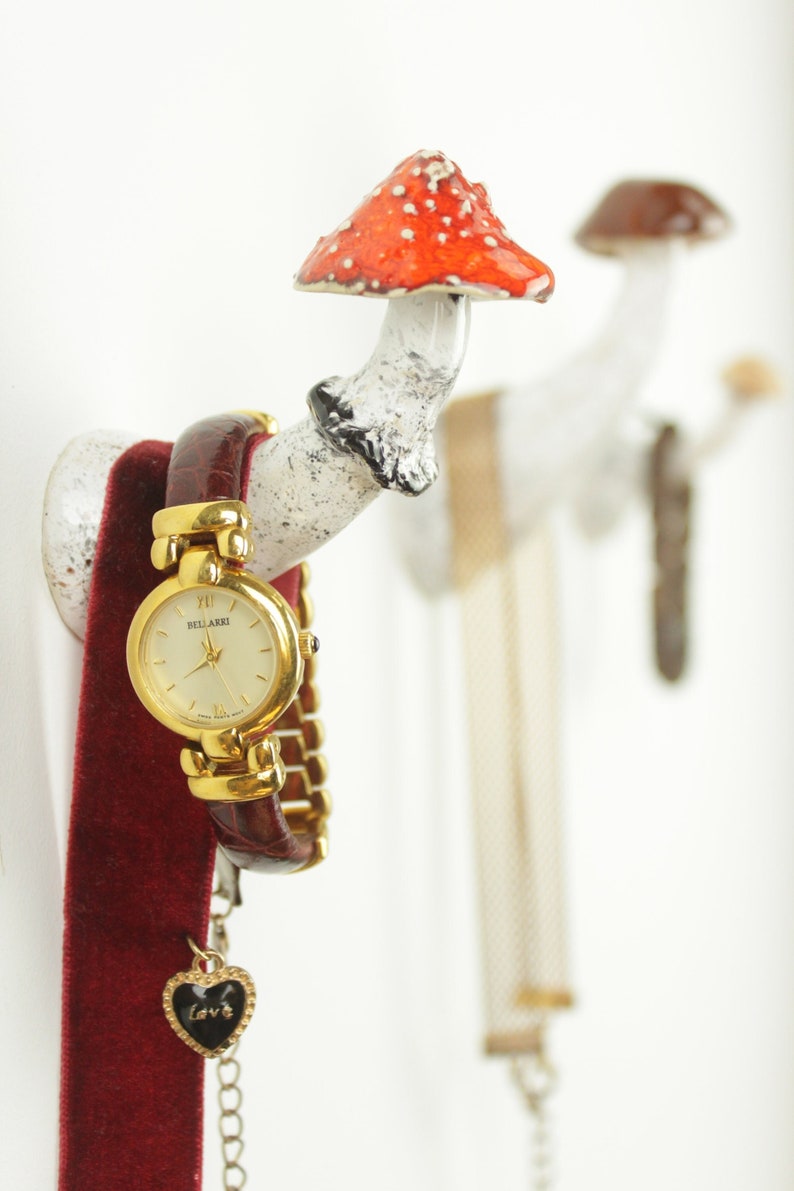 May include: A white ceramic mushroom-shaped hook with a red and white mushroom cap. A gold watch with a brown leather band and a gold chain with a heart charm hangs from the hook. A gold chain necklace hangs from a second hook.