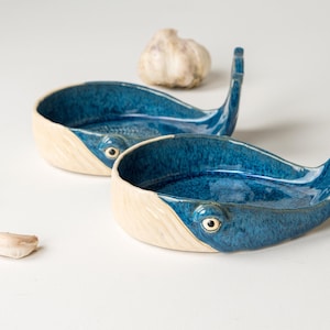 May include: Two ceramic whale-shaped spoon rests. The interior is a glossy blue, contrasting with the off-white exterior. Each whale has a tail fin and a small eye detail. Garlic cloves and a bulb of garlic are also present.