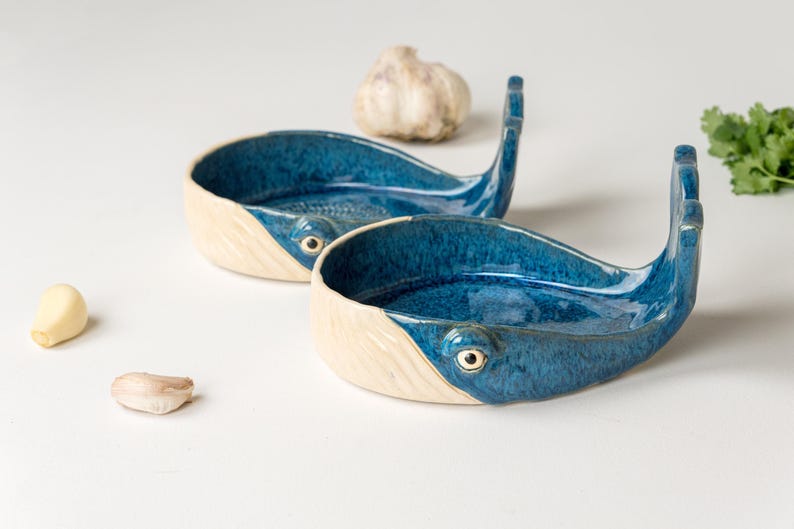 May include: Two ceramic whale-shaped spoon rests. The interior is a glossy blue, contrasting with the off-white exterior. Each whale has a tail fin and a small eye detail. Garlic cloves and a bulb of garlic are also present.
