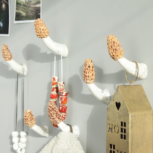 May include: Five white ceramic mushroom-shaped hooks mounted on a wall. The hooks have brown tops and are decorated with a textured pattern. Two necklaces and a string of red beads hang from the hooks. A gold house-shaped ornament hangs from one of the hooks.