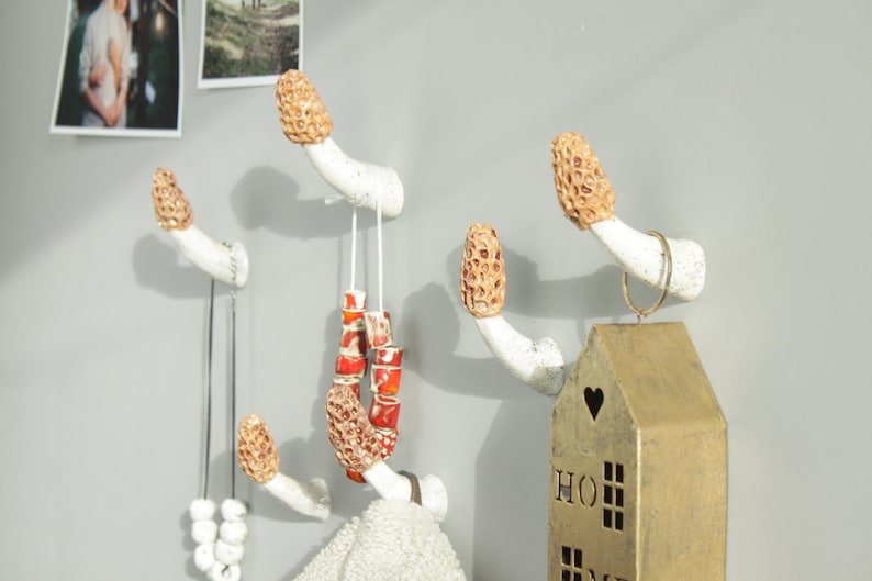 May include: Five white ceramic mushroom-shaped hooks mounted on a wall. The hooks have brown tops and are decorated with a textured pattern. Two necklaces and a string of red beads hang from the hooks. A gold house-shaped ornament hangs from one of the hooks.