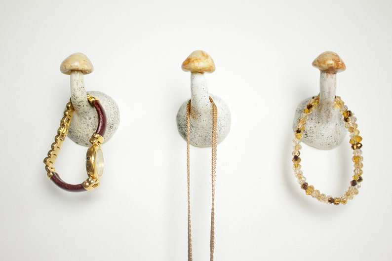 Puede incluir: Tres ganchos de cer&aacute;mica con forma de hongo en una pared blanca. Los ganchos est&aacute;n decorados con un esmalte blanco y marr&oacute;n moteado. Un reloj, un collar y un brazalete de cuentas cuelgan de los ganchos.