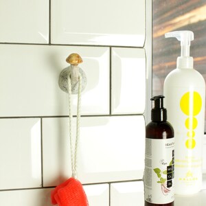 May include: White ceramic mushroom-shaped hook with a white rope hanging from it. A red and white bath sponge is hanging from the rope. Two bottles of body wash are on a shelf in the background.