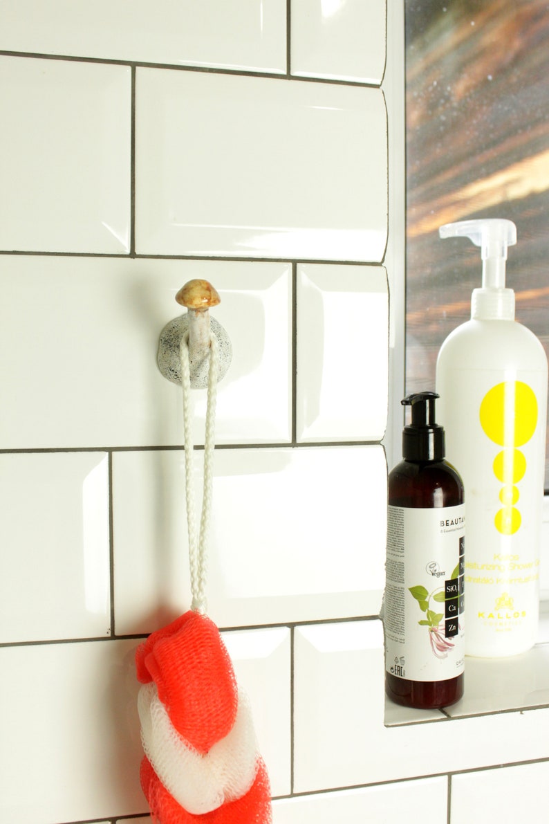 May include: White ceramic mushroom-shaped hook with a white rope hanging from it. A red and white bath sponge is hanging from the rope. Two bottles of body wash are on a shelf in the background.