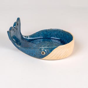 May include: A ceramic whale-shaped dish with a textured, speckled blue body and a cream-colored tail. The dish features a detailed eye and a decorative pattern inside the bowl. The whale's tail has three fin-like protrusions.