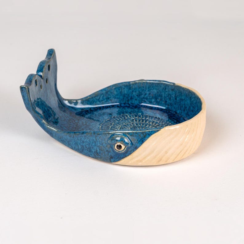 May include: A ceramic whale-shaped dish with a textured, speckled blue body and a cream-colored tail. The dish features a detailed eye and a decorative pattern inside the bowl. The whale's tail has three fin-like protrusions.
