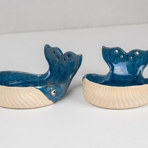 May include: Two ceramic whale-shaped dishes. The whale tails are a deep blue with three holes, and the bodies are a creamy white. The dishes have a glossy finish and are designed to hold small items. The whale's eye is visible.