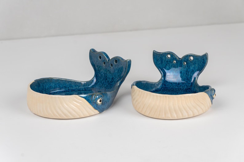 May include: Two ceramic whale-shaped dishes. The whale tails are a deep blue with three holes, and the bodies are a creamy white. The dishes have a glossy finish and are designed to hold small items. The whale's eye is visible.