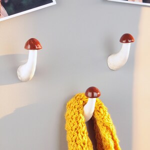 May include: Two white ceramic mushroom-shaped hooks with brown tops mounted on a gray wall. A yellow knitted scarf is hanging on one of the hooks.