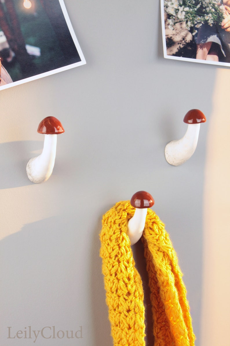 May include: Two white ceramic mushroom-shaped hooks with brown tops mounted on a gray wall. A yellow knitted scarf is hanging on one of the hooks.