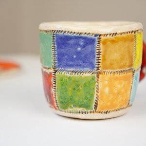 K&ouml;nnte beinhalten: Eine Keramiktasse mit Patchwork-Design, das aus Quadraten in Blau, Gr&uuml;n, Gelb und Orange besteht. Die Tasse hat einen Henkel und ist mit braunen N&auml;hten um jedes Quadrat verziert.
