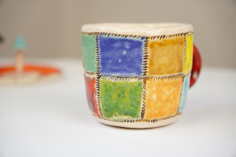 K&ouml;nnte beinhalten: Eine Keramiktasse mit Patchwork-Design, das aus Quadraten in Blau, Gr&uuml;n, Gelb und Orange besteht. Die Tasse hat einen Henkel und ist mit braunen N&auml;hten um jedes Quadrat verziert.