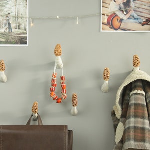 May include: Six ceramic mushroom-shaped hooks on a white wall. A brown leather backpack hangs on one hook, a plaid jacket hangs on another, and a string of red beads hangs on a third hook.