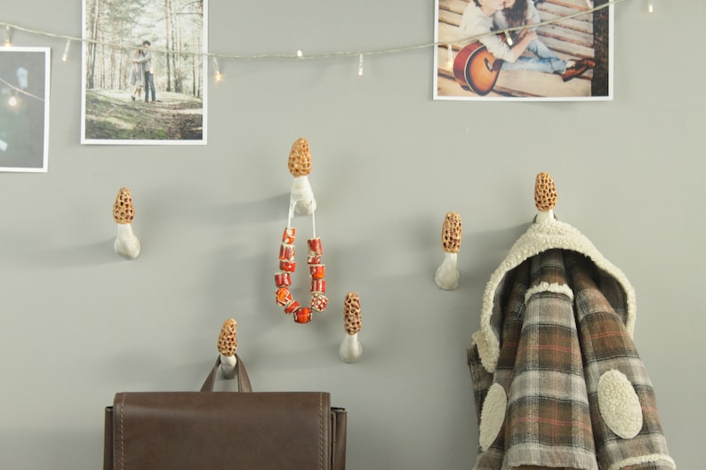 May include: Six ceramic mushroom-shaped hooks on a white wall. A brown leather backpack hangs on one hook, a plaid jacket hangs on another, and a string of red beads hangs on a third hook.