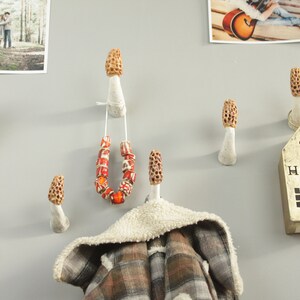 May include: A wall with six white ceramic mushroom-shaped hooks. The hooks are decorated with brown and orange details. A necklace, a scarf, and a small wooden sign that says "HOME" are hanging on the hooks.