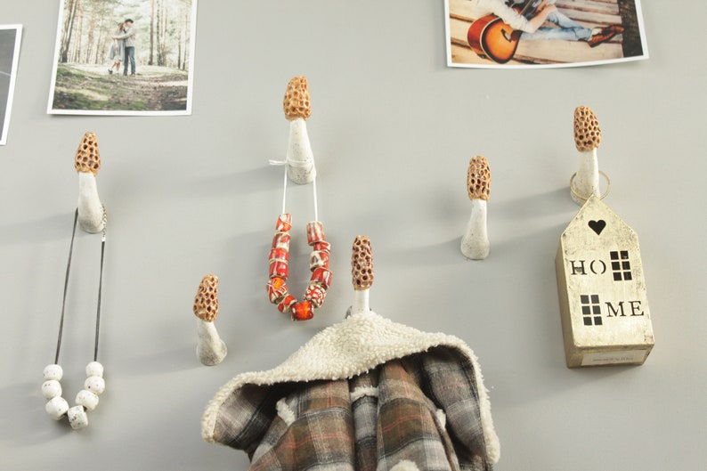 May include: A wall with six white ceramic mushroom-shaped hooks. The hooks are decorated with brown and orange details. A necklace, a scarf, and a small wooden sign that says "HOME" are hanging on the hooks.