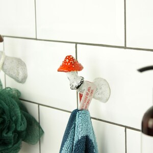 May include: Two white ceramic mushroom hooks with red and brown accents mounted on a white tiled wall. A green bath sponge hangs from one hook and a blue towel hangs from the other. A brown bottle of hand soap with a black pump is on the right side of the image.