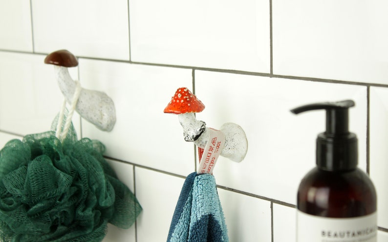 May include: Two white ceramic mushroom hooks with red and brown accents mounted on a white tiled wall. A green bath sponge hangs from one hook and a blue towel hangs from the other. A brown bottle of hand soap with a black pump is on the right side of the image.