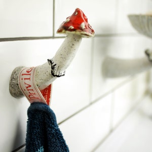 In the photo there is a wall hook in the shape of a fly agaric. He has a white leg with black pollination, a cute skirt with a black border and a bright red-orange hat. There is a towel hanging from a hook growing from the wall.