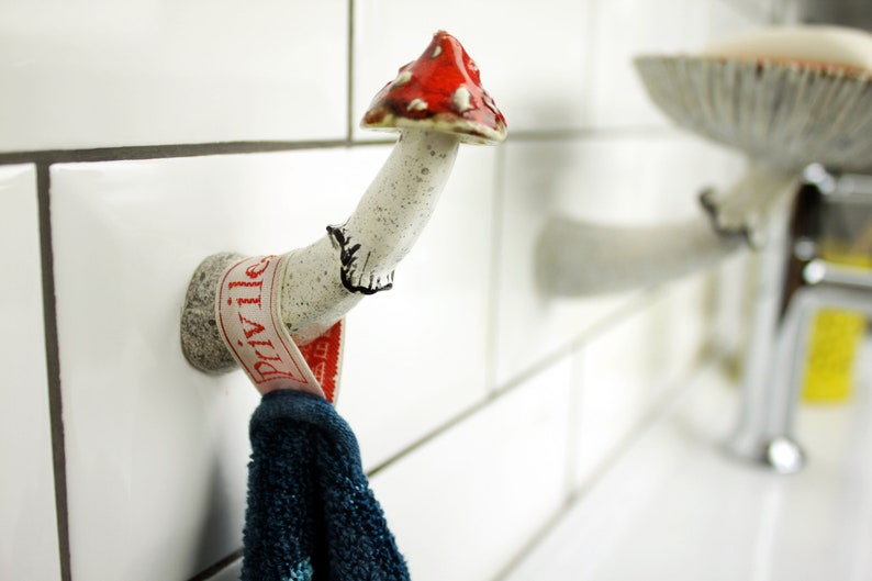 In the photo there is a wall hook in the shape of a fly agaric. He has a white leg with black pollination, a cute skirt with a black border and a bright red-orange hat. There is a towel hanging from a hook growing from the wall.