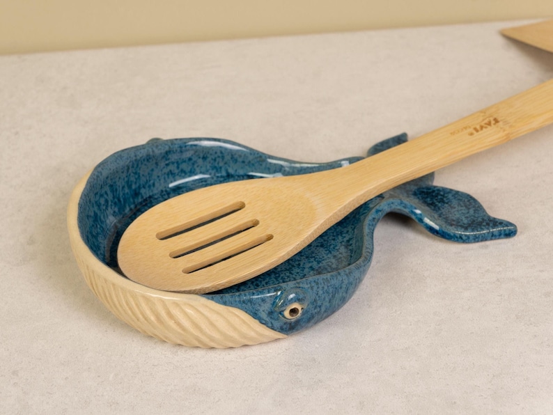 May include: A blue and white ceramic whale-shaped spoon rest with a wooden spoon resting inside. The spoon has a slotted design and the text "ZAVIT" is printed on the handle.