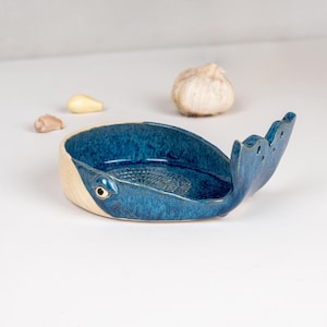 May include: A ceramic whale-shaped garlic grater with a blue glazed interior and a natural beige exterior. The whale's tail is raised, and the grater has a textured surface. Garlic cloves are visible in the background.