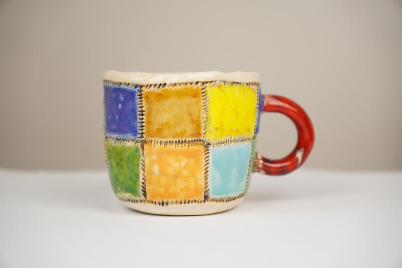 May include: A ceramic mug with a patchwork design in shades of blue, green, yellow, and orange. The mug has a red handle.