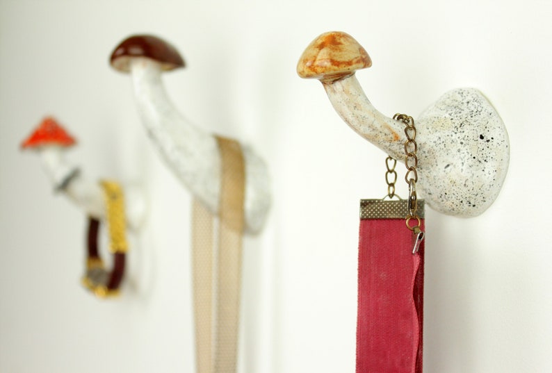 May include: Two white ceramic mushroom-shaped hooks with a brown and orange mushroom cap. One hook has a gold chain with a red ribbon hanging from it. The other hook has a gold chain with a brown leather strap hanging from it.