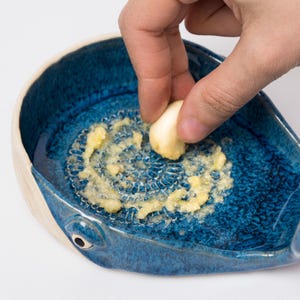 May include: A blue ceramic garlic grater in the shape of a whale. The grater has a textured surface and is filled with grated garlic. A hand holds a garlic clove over the grater.