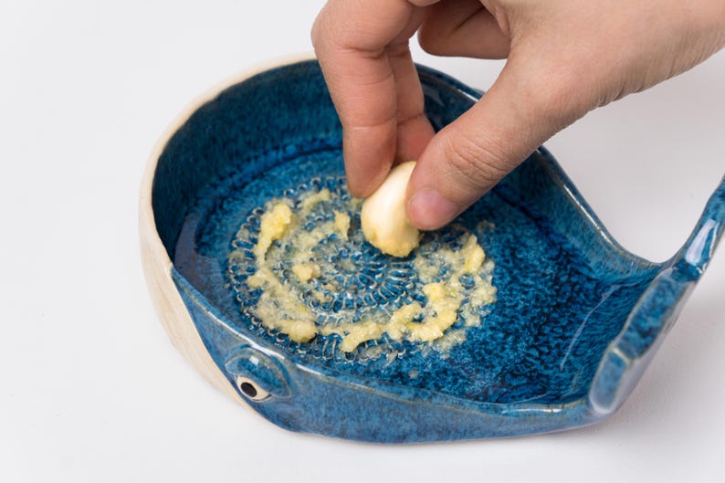 May include: A blue ceramic garlic grater in the shape of a whale. The grater has a textured surface and is filled with grated garlic. A hand holds a garlic clove over the grater.