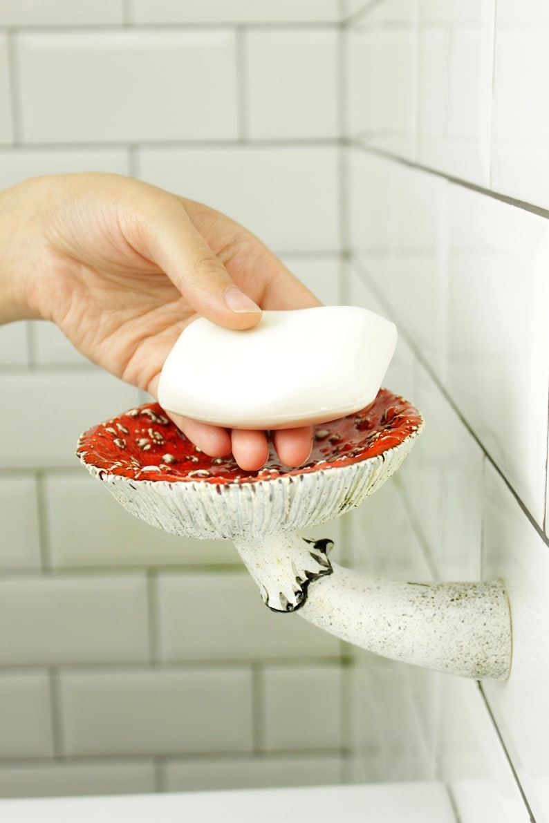 May include: A white ceramic soap dish shaped like a red mushroom with a white stem. The soap dish is mounted on a white tiled wall.