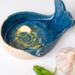 May include: A blue ceramic garlic grater in the shape of a whale, with a section of grated garlic. A whole garlic bulb and a basil leaf are also present. The grater has a white rim and a textured surface.