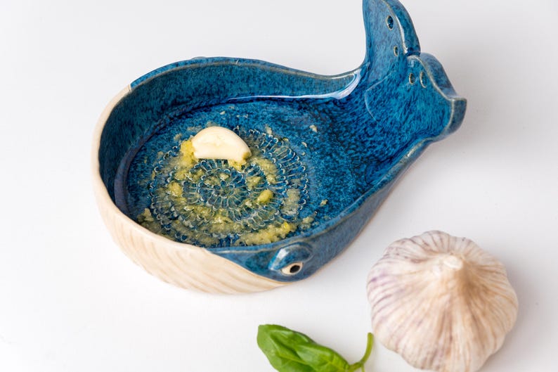 May include: A blue ceramic garlic grater in the shape of a whale, with a section of grated garlic. A whole garlic bulb and a basil leaf are also present. The grater has a white rim and a textured surface.