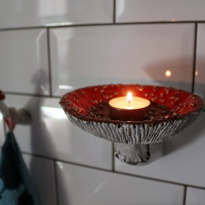 In the twilight in the bathroom, a candle was lit, which stands in the hat of the fly agaric shelf. The gentle light from the flame illuminates the relief of the hat.In the background you can see a fly agaric hook with a towel on it.