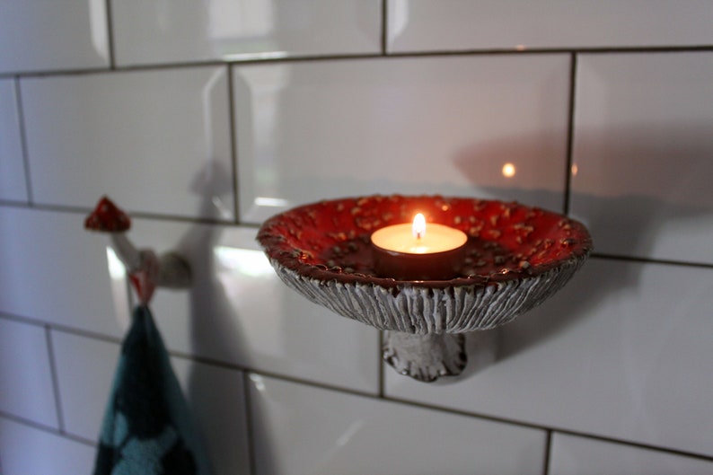 In the twilight in the bathroom, a candle was lit, which stands in the hat of the fly agaric shelf. The gentle light from the flame illuminates the relief of the hat.In the background you can see a fly agaric hook with a towel on it.