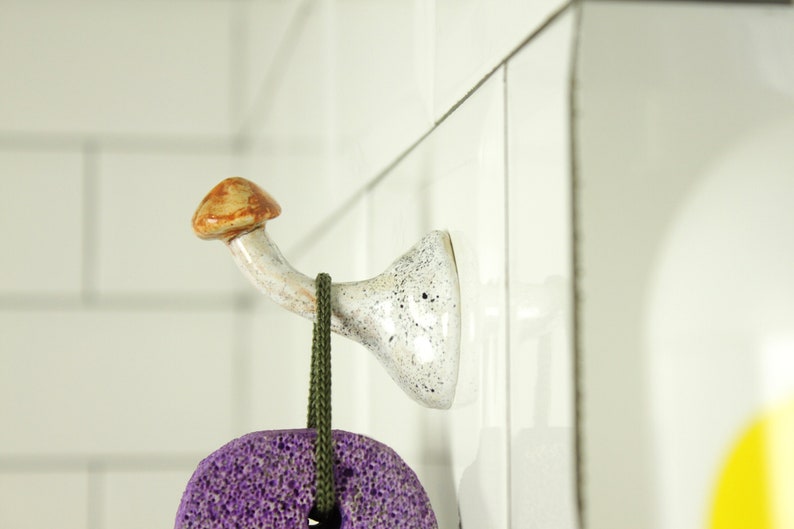 May include: A white ceramic mushroom-shaped hook with brown accents mounted on a white tiled wall. A purple and green scrub brush hangs from the hook.