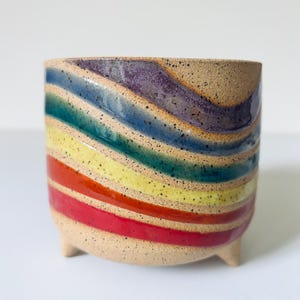 May include: A cylindrical ceramic planter with three small feet, featuring a speckled beige base and diagonal stripes in purple, blue, teal, yellow, orange, and red. The surface has a glossy finish over the colorful bands, contrasting with the textured, speckled clay body visible between the stripes.