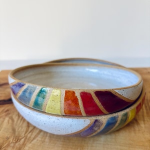 May include: Two stacked ceramic bowls with a speckled white finish and a vibrant rainbow-colored band painted around the exterior rim. The colorful stripes feature shades of purple, blue, teal, yellow, orange, and red on a light clay base.