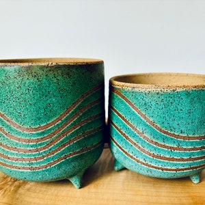 May include: Two ceramic planters with a speckled turquoise glaze and brown wavy lines. The planters have a rounded shape and sit on three small feet. The rims and interior are a natural tan color. The planters are suitable for indoor or outdoor use.