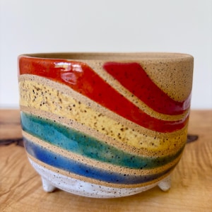 May include: A small, round, speckled ceramic planter with three short feet. The exterior features horizontal, wavy bands of glossy red, yellow, teal, and blue glaze over a tan, speckled clay base, with a white glazed bottom section.