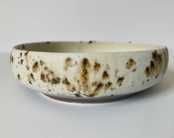 Könnte beinhalten: Eine Keramikschale mit gesprenkeltem Design. Die Schale hat einen cremefarbenen Innen- und Außenbereich mit braunen Sprenkeln. Die Schale ist rund und hat eine breite, flache Form.