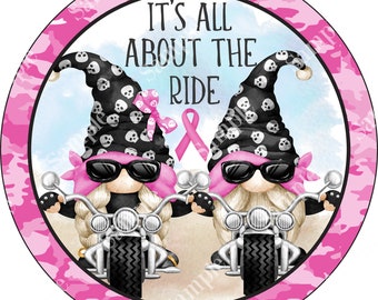 Puede incluir: Dos gnomos montando en motocicletas con bandanas rosas y gafas de sol. Los gnomos llevan sombreros negros y blancos con calaveras. El fondo es de camuflaje rosa con una cinta rosa y el texto "It's All About The Ride".