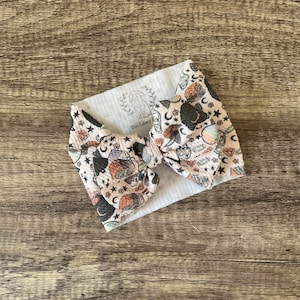 May include: A pink and black hair bow with a witchy print featuring stars, moons, potions, and other magical elements. The bow is tied with a white ribbon and has the text "The Lovey" on the ribbon.