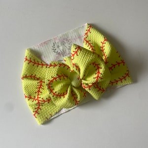 May include: A yellow and red softball-themed hair bow. The bow is made of a soft, textured fabric and features a red stitching pattern that resembles a softball.