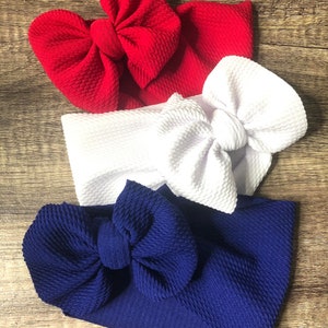 May include: Three fabric headbands in red, white, and blue. Each headband features a large bow on top. The headbands are made of a textured material and are arranged on a wooden surface.