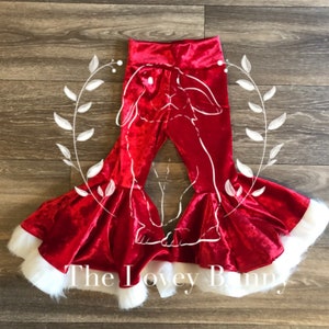 Può includere: Pantaloni a zampa di elefante in velluto rosso con finiture in ecopelliccia bianca sull'orlo. I pantaloni hanno una vita alta e un design a contorno bianco. Il testo "The Lovey Bunny" è nella parte inferiore dell'immagine.