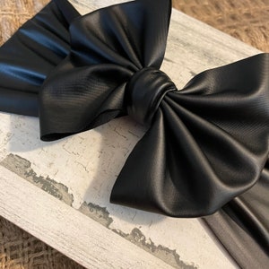 May include: A large black faux leather bow headband. The headband is made of a soft, pliable material and has a large, dramatic bow.