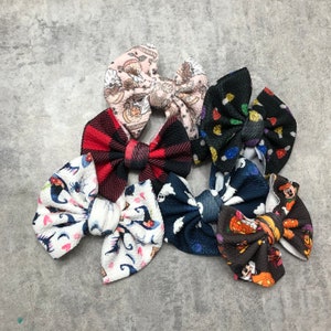 May include: A set of 7 Halloween-themed hair bows. The bows are made of fabric and feature various patterns, including ghosts, pumpkins, and bats. The bows are in a variety of colors, including black, white, orange, and purple.